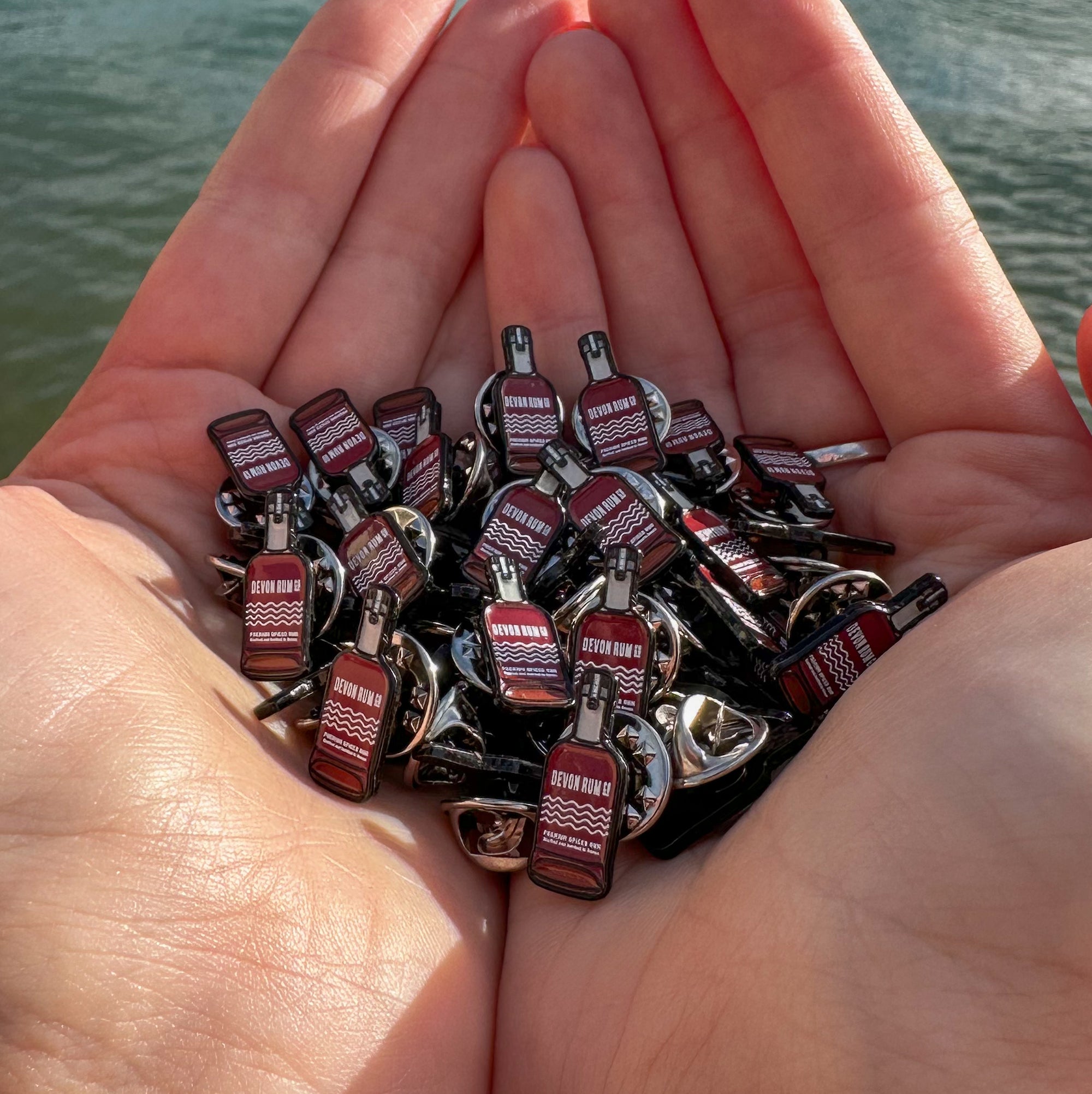 Small Enamel Devon Rum Co Spiced Rum Bottle Pins in Hands Over the Water in Salcombe, Devon