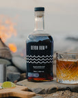 Devon Rum Co Black Spiced Rum bottle and a ginger ale cocktail on the beach with a bonfire behind.