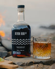 Devon Rum Co Black Spiced Rum bottle and a ginger ale cocktail on the beach with a bonfire behind.