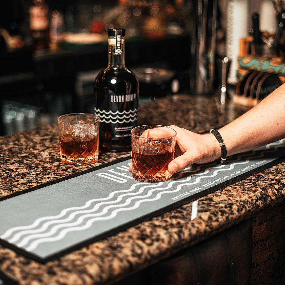 A bar runner with a white wave design, placed on a countertop with a person holding a glass of rum and a bottle in the background.