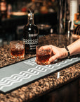 A bar runner with a white wave design, placed on a countertop with a person holding a glass of rum and a bottle in the background.