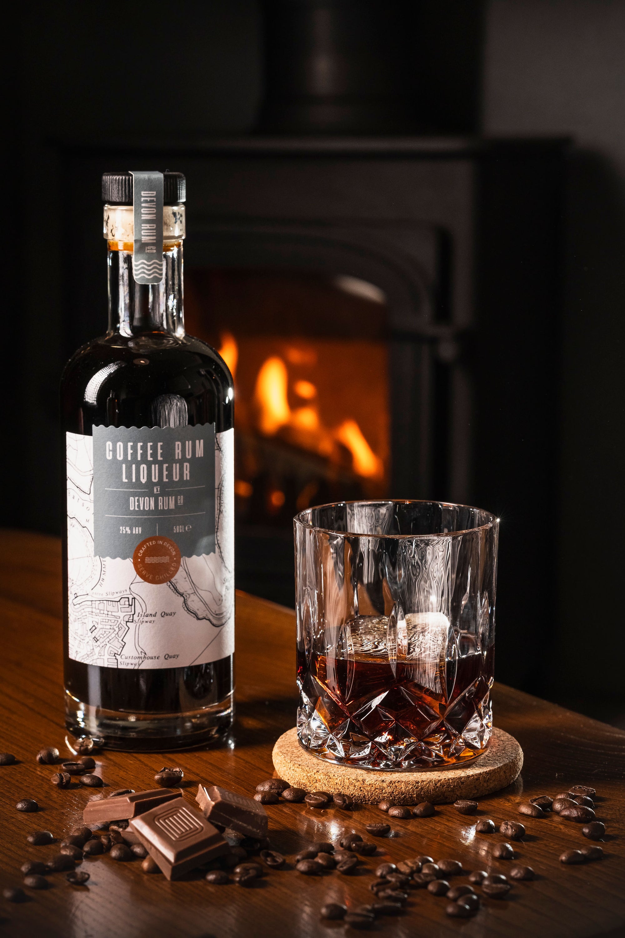 Glass of Devon Rum Co Coffee Rum Liqueur with a bottle and coffee beans in front of a fireplace.