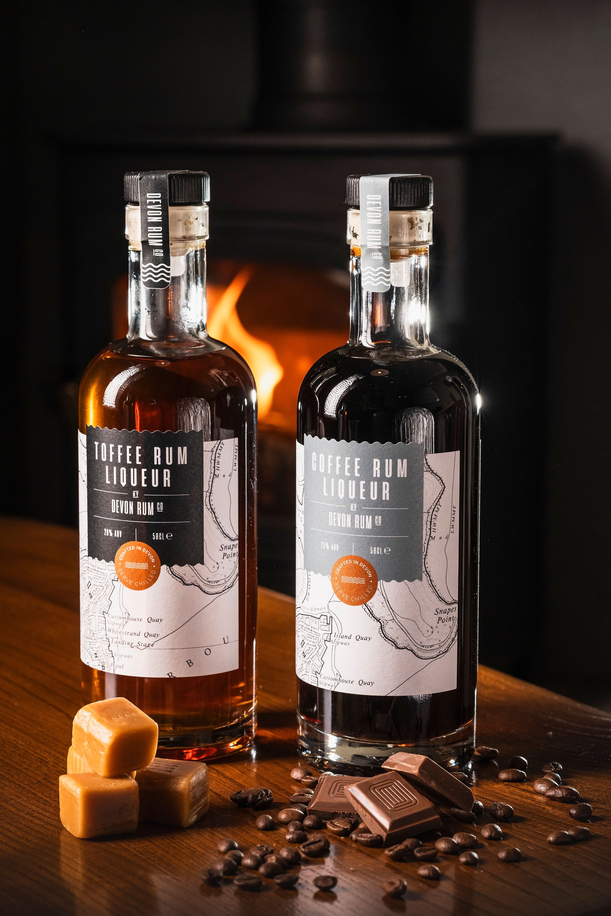 Two bottles of Toffee & Coffee Rum Liqueur on a wooden surface with coffee beans and chocolate, fireplace in the background.
