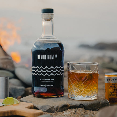 Devon Rum Co Black Spiced Rum bottle and a ginger ale cocktail on the beach with a bonfire behind.